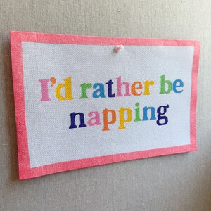 Pode incluir: Uma tela retangular de ponto de agulha com o texto "I'd rather be napping" em letras multicoloridas. A tela tem um fundo de grade branca e é contornada por uma moldura de fita rosa sólida, fixada no centro superior com um pequeno alfinete branco.