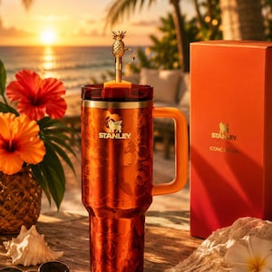 May include: A metallic orange Stanley tumbler with a handle, a dark lid, and a gold pineapple-shaped straw topper. The tumbler features a subtle floral pattern and the Stanley logo. Beside it stands a matching orange box labeled Stanley Iconic Edition.