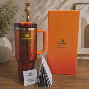 May include: A red and orange Stanley Quencher H2.0 tumbler with a floral pattern, a gold pineapple straw topper, and an orange handle. Beside it stands a tall orange box labeled Stanley Karol G, along with a small white product information booklet.