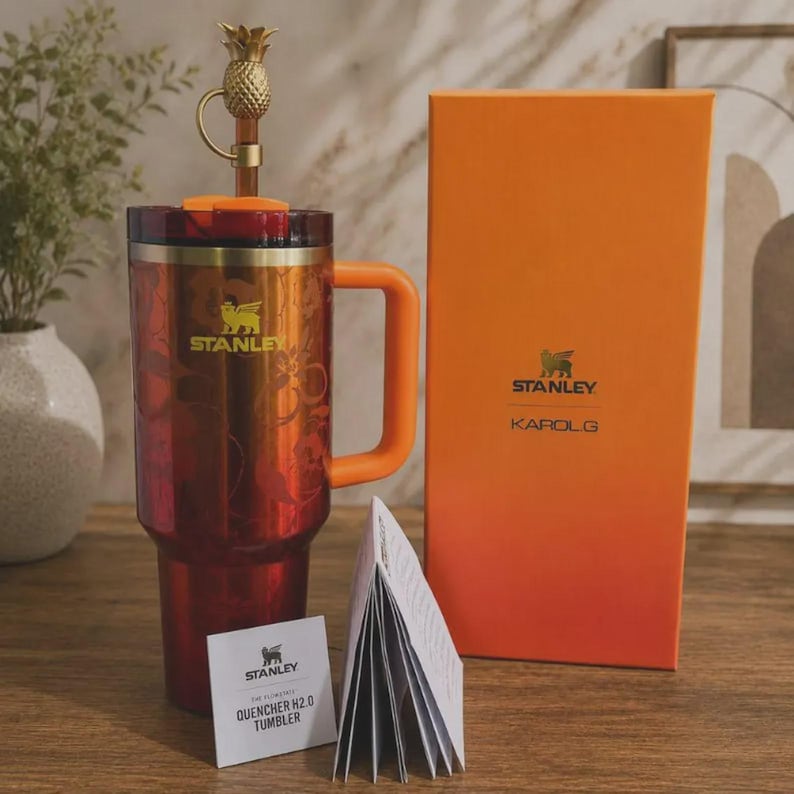 May include: A red and orange Stanley Quencher H2.0 tumbler with a floral pattern, a gold pineapple straw topper, and an orange handle. Beside it stands a tall orange box labeled Stanley Karol G, along with a small white product information booklet.