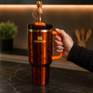 May include: A copper-colored Stanley tumbler with a marbled finish, featuring an orange handle and a dark lid. A gold-toned metal pineapple topper is attached to the straw. The Stanley logo is printed in gold on the front of the insulated cup.