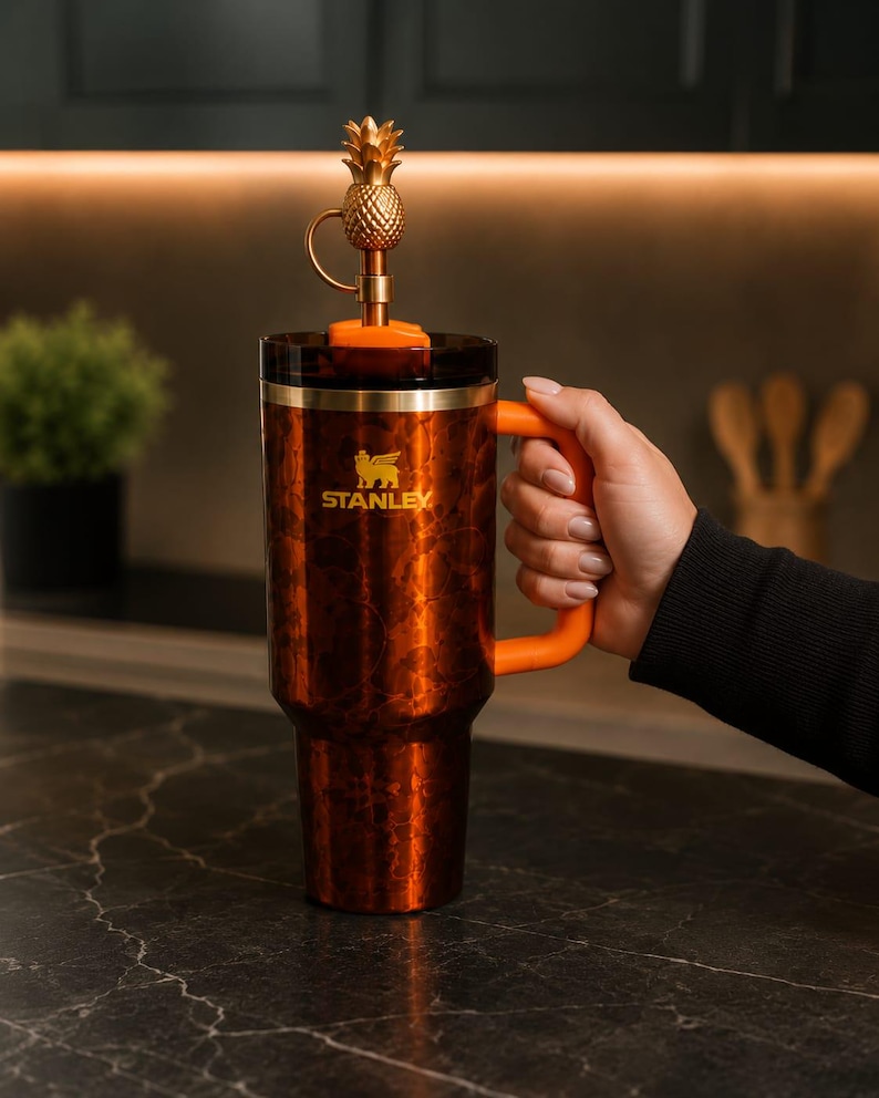 May include: A copper-colored Stanley tumbler with a marbled finish, featuring an orange handle and a dark lid. A gold-toned metal pineapple topper is attached to the straw. The Stanley logo is printed in gold on the front of the insulated cup.