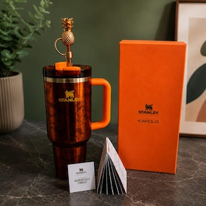 May include: A brown patterned Stanley Quencher H2.0 tumbler with an orange handle, a gold pineapple-shaped straw topper, and a matching orange box labeled Stanley Karol G. A small white product information card is placed in front of the tumbler.