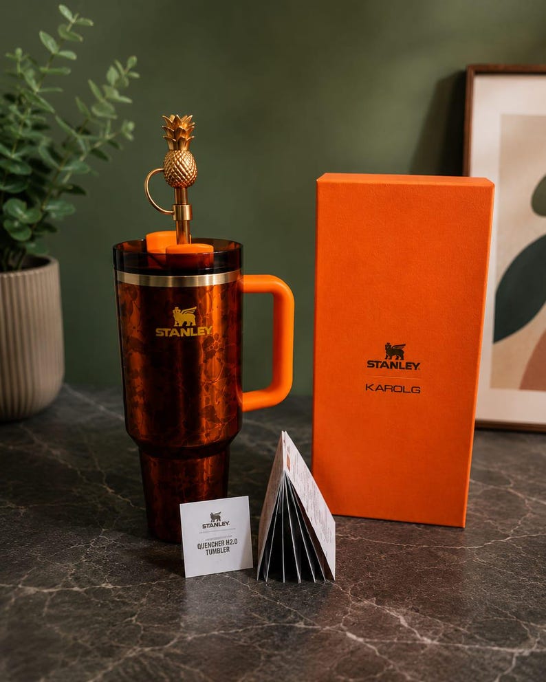 May include: A brown patterned Stanley Quencher H2.0 tumbler with an orange handle, a gold pineapple-shaped straw topper, and a matching orange box labeled Stanley Karol G. A small white product information card is placed in front of the tumbler.