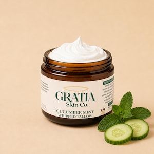 May include: A brown glass jar filled with white whipped tallow, topped with a peaked swirl. The label reads Gratia Skin Co. Cucumber Mint Whipped Tallow. Beside the jar are two fresh cucumber slices and a sprig of green mint leaves.