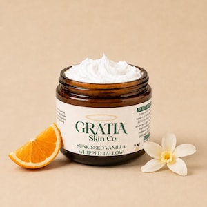 Op de afbeelding: Een bruine glazen pot gevuld met witte opgeklopte talk, met een etiket met de tekst Gratia Skin Co. Sunkissed Vanilla Whipped Tallow. Een schijfje sinaasappel ligt links en een witte vanillebloem ligt rechts van de pot.
