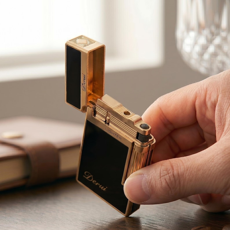 May include: A gold-toned metal lighter with a rectangular shape and black panels. The lid is open, revealing the internal mechanism. The word Derui is printed on the front and inside the lid, with the word LIGHTER printed on the top edge.