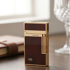 May include: A rectangular pocket lighter featuring a polished gold-tone metal frame and dark brown tortoiseshell-patterned panels. The word LIGHTER is printed in small black letters near the top, and a stylized letter D logo is visible at the bottom.