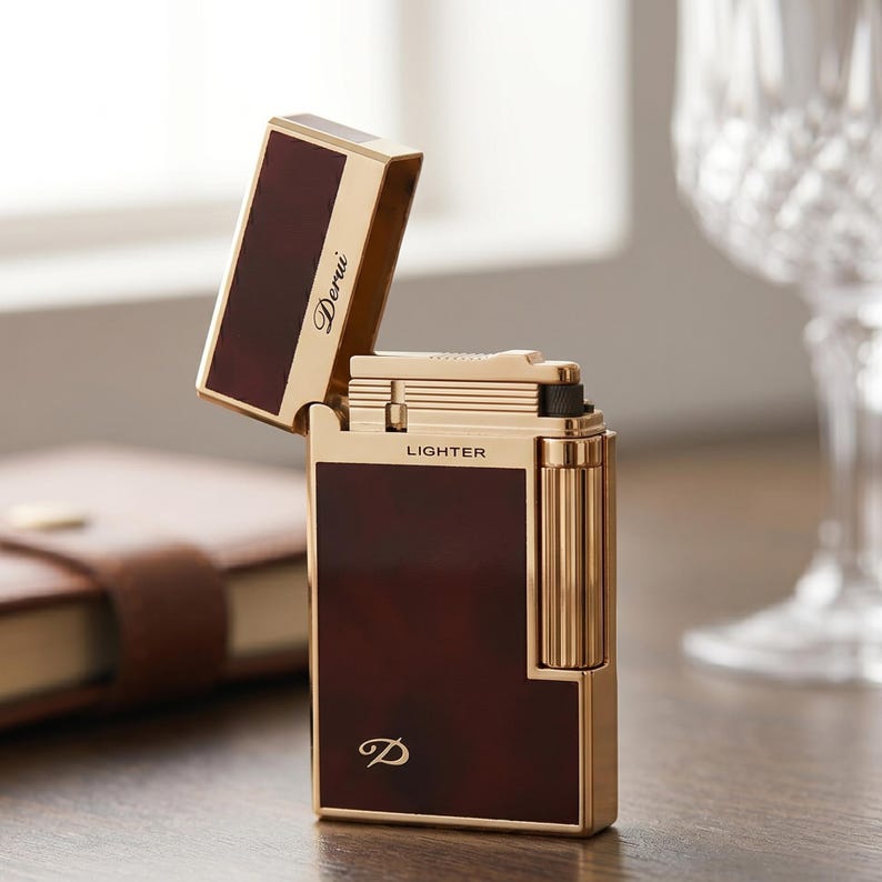 May include: A gold-toned metal lighter with dark brown rectangular panels. The open lid displays the script text Derui, while the body features the word LIGHTER in block letters and a stylized D logo at the bottom. The design includes a vertical roller.