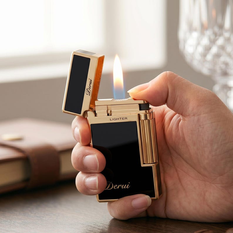 May include: A gold-toned metal lighter with black rectangular panels on the front and lid. The word Derui is printed in white script on the lid and body, with the word LIGHTER printed in block letters. A small blue and orange flame is visible.