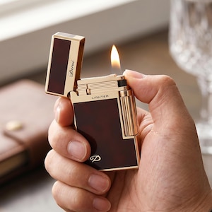 May include: A gold-toned metal lighter with a dark brown tortoiseshell-patterned inlay. The lighter is open with a visible flame, featuring the word "LIGHTER" printed on the top edge and "Derui" on the lid, with a stylized "D" logo at the base.