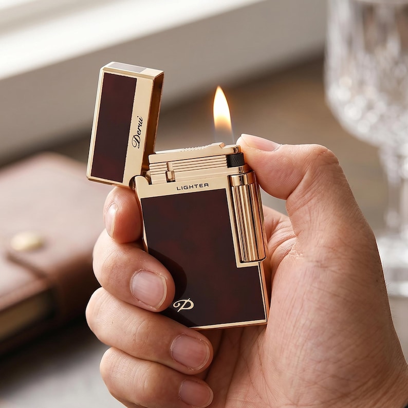 May include: A gold-toned metal lighter with a dark brown tortoiseshell-patterned inlay. The lighter is open with a visible flame, featuring the word "LIGHTER" printed on the top edge and "Derui" on the lid, with a stylized "D" logo at the base.