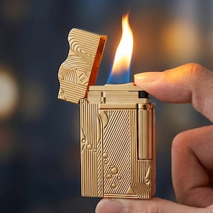 以下が含まれることがあります： 蓋が開いた状態で炎が出ている金色の金属製ライター。長方形の本体には、斜めの線と小さな円形の模様が施された質感のある表面があります。ライターは直立した状態で保持されており、その金属的な仕上げとデザインが示されています。