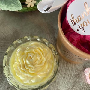 Pode incluir: Uma vela amarelo-pálido em forma de rosa, dentro de um recipiente de vidro transparente facetado. Ao lado, uma caixa de presente cilíndrica de papelão preenchida com papel de seda rosa vibrante, amarrada com uma fita branca e um adesivo de agradecimento.