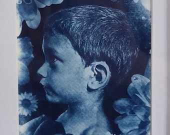 May include: A cyanotype print featuring a profile portrait of a child surrounded by floral elements, presented in a rectangular wooden frame with a white mat. The artwork uses monochromatic blue tones to depict the subject and surrounding flowers.