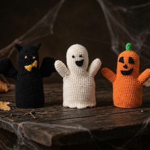 Può includere: Tre marionette da dito all'uncinetto composte da un pipistrello nero con dettagli gialli, un fantasma bianco con occhi e bocca neri e una zucca arancione con picciolo verde e tratti del viso neri, disposte in fila.