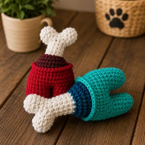May include: Two crocheted toys shaped like meat on the bone. One toy features a red base with a dark maroon section and a white bone top. The second toy has a teal base with a dark blue section and a white bone end, both crafted with yarn.