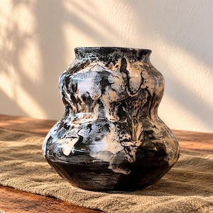 May include: A ceramic vase with a bulbous, double-gourd shape featuring a high-gloss glaze. The surface displays a marbled, abstract pattern of deep black and creamy white colours that swirl together across the entire exterior body.