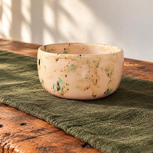 Può includere: Una ciotola in ceramica rotonda con una smaltatura di base rosa pallido, caratterizzata da un motivo irregolare a macchie nei toni del marrone scuro, beige e verde chiaro. La ciotola ha una finitura liscia e lucida e un bordo irregolare.