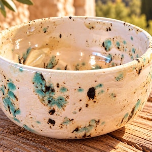 Può includere: Una ciotola in ceramica rotonda con una smaltatura lucida color bianco sporco, caratterizzata da macchie irregolari marrone scuro e turchese brillante sparse sulle superfici interne ed esterne. La ciotola ha un bordo liscio e una base arrotondata.