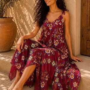 Puede incluir: Un vestido midi sin mangas en un tono burdeos profundo con un vibrante estampado floral y geométrico en tonos blanco, azul, amarillo y verde. El vestido tiene tirantes finos para anudar, escote en V y un bajo asimétrico en punta.