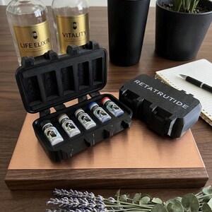 May include: Two black rectangular storage cases with foam inserts, one open and one closed with the text RETATRUTIDE printed on top. The open case holds five small glass vials with white labels and colored caps, arranged in a row on a wooden surface.