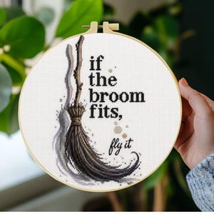 Könnte beinhalten: Ein runder Stickrahmen mit einem Kreuzstichmotiv eines braunen Besens, dem schwarzen Text "if the broom fits, fly it" und kleinen grauen Sternakzenten auf weißem Stoff, eingefasst in einen hellen Holzrahmen.