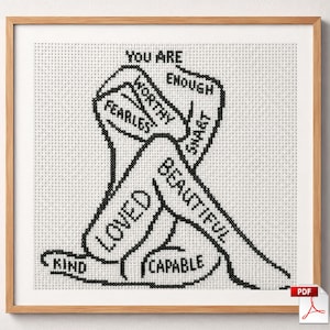Puede incluir: Un patrón de punto de cruz en un marco de madera clara que muestra el contorno negro de una figura sentada. La figura está compuesta por las palabras: You Are Enough, Worthy, Fearless, Smart, Loved, Beautiful, Kind y Capable, bordadas en tela blanca.