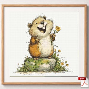 May include: A cross-stitch pattern featuring a cheerful, brown and cream-colored groundhog standing on a mossy, green rock. The groundhog is holding a small yellow flower in one hand. The design is framed in a light-colored wooden frame.