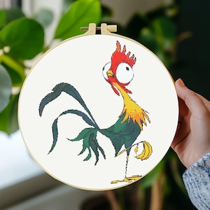 May include: A circular wooden embroidery hoop containing a cross-stitch pattern of a cartoon rooster with large white eyes, a bright red comb, a yellow neck, and dark green tail feathers, set against a plain white fabric background.