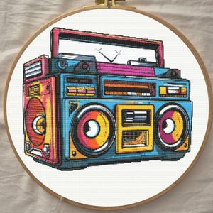 Puede incluir: Un bordado de punto de cruz que presenta un radiocassette de estilo pixel-art vibrante. El diseño muestra una paleta de colores azul, rosa y amarillo con dos grandes altavoces circulares, un asa superior y una antena, todo enmarcado en un bastidor de madera circular.