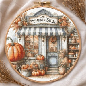May include: A cross-stitch embroidery in a round wooden hoop depicting a storefront with the text PUMPKIN STORE. The scene features a blue and white striped awning, a large orange pumpkin in the foreground, and shelves filled with various pumpkins.