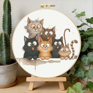 May include: A circular wooden embroidery hoop containing a cross-stitch pattern of six cartoon cats with wide eyes. The cats are depicted in shades of gray, brown, black, and orange, arranged in a group on a white fabric background.