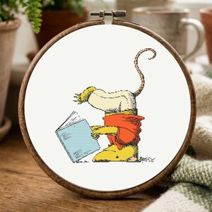 May include: A circular wooden embroidery hoop framing a cross-stitch design of a stylized creature with a long tail, wearing a red shirt and yellow pants, while holding and reading an open light blue book against a plain white background.