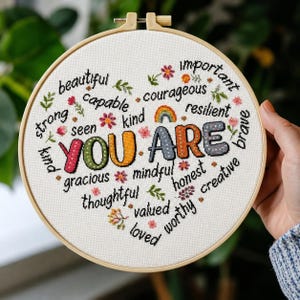 Può includere: Un telaio da ricamo circolare in legno contenente tessuto bianco con un disegno a punto croce. Il testo YOU ARE è centrato in grandi lettere colorate e decorate, circondato da parole nere più piccole come beautiful, capable e resilient.