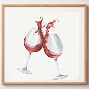 May include: A cross-stitch embroidery pattern featuring two clear wine glasses clinking together, with deep red wine splashing out of the rims. The design is framed in a light-colored wooden square frame against a white background.