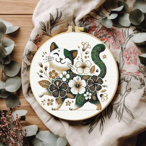 May include: A circular wooden embroidery hoop containing a cross-stitch pattern of a smiling cat with green and white fur, surrounded by detailed floral motifs in shades of dark green, brown, and white, set against a plain white fabric background.