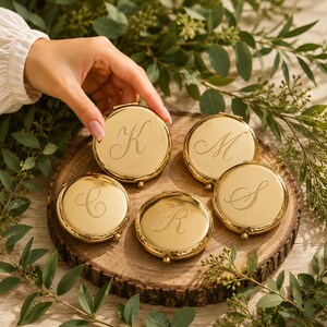 May include: Five round, gold-toned metal compact mirrors, each featuring a single engraved cursive letter. The letters K, M, C, R, and S are centered on the polished, reflective surfaces of the circular cases, which have a hinged closure.