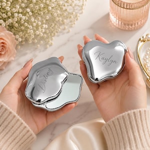 May include: Two silver-toned heart-shaped compact mirrors with a polished metallic finish. One mirror is closed and engraved with the name Kaylyn, while the other is open, revealing the reflective surface and engraved with the name Destiny.