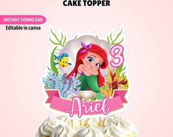 May include: A cake topper featuring a cartoon mermaid with red hair, a yellow fish, and a red crab inside a shell. The design includes green seaweed, blue coral, a pink number 3, and a pink banner with the name Ariel in white script text.