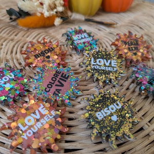 Peut inclure: Une collection de badges en résine en forme de soleil avec des finitions pailletées et diverses phrases dont LOVE YOURSELF, BISOU, COLORE TA VIE et PERLE RARE. Chaque badge a un contour pointu en étoile et affiche du texte avec des petits cœurs.