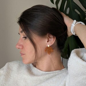 Peut inclure: Une paire de boucles d'oreilles en forme de feuille d'érable, translucides et de couleur ambre, avec une surface texturée et une petite attache en métal doré sur le dessus, portées au lobe.