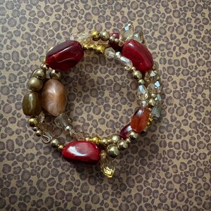 May include: A multi-strand wrap bracelet featuring a mix of smooth red, peach, and brown oval beads interspersed with metallic gold spherical beads and clear faceted crystal accents. The design consists of several circular layers of beads.