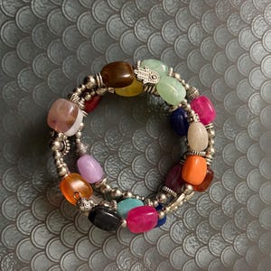 May include: A multi-strand wrap bracelet featuring smooth, polished, multicolored beads in shades of brown, yellow, green, pink, blue, orange, and black, interspersed with small silver-toned metal beads and a silver-toned hamsa hand charm.