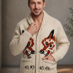 May include: A cream-colored knit cardigan featuring a shawl collar, long sleeves, and two front pockets. The garment is decorated with a repeating intarsia pattern of stylized fox faces in shades of orange, black, and white, with ribbed cuffs and hem.