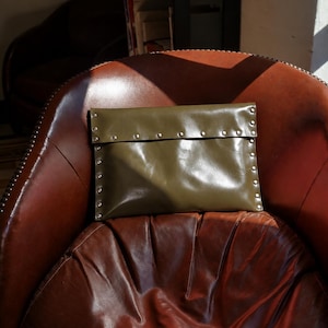 Può includere: Una pochette rettangolare color verde oliva con design a patta ripiegabile. I bordi della borsa e della patta sono impreziositi da una serie di piccoli rivetti metallici color argento, distanziati uniformemente sulla superficie liscia.