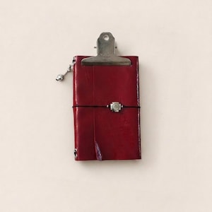 Puede incluir: Un cuaderno de cuero rojo oscuro asegurado con una banda elástica negra que presenta un dije cuadrado de tono plateado en el centro. Un clip de metal plateado está sujeto al borde superior y un pequeño abalorio plateado cuelga del lateral.