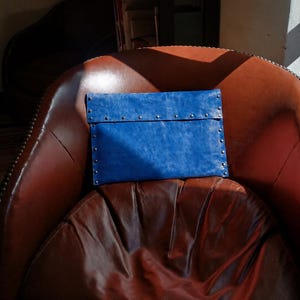 May include: A rectangular blue clutch bag with a distressed leather-like texture, featuring a fold-over flap secured with a row of small silver-toned metal studs along the edges and the center seam, creating a structured and polished appearance.