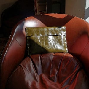 Op de afbeelding: Een rechthoekige olijfgroene clutch met een glanzende, getextureerde afwerking. De tas heeft een overslagflap die is vastgezet met een rij kleine, zilverkleurige metalen studs langs de randen en de horizontale naad van de sluiting.