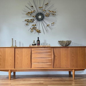 Op de afbeelding: Een lang houten dressoir met aan weerszijden twee schuifdeuren en vier centrale lades. Het meubelstuk heeft taps toelopende poten en een lichtbruine houtnerf. Bovenop staan twee koperen kandelaars, een houten dienblad met flessen en een schaal.